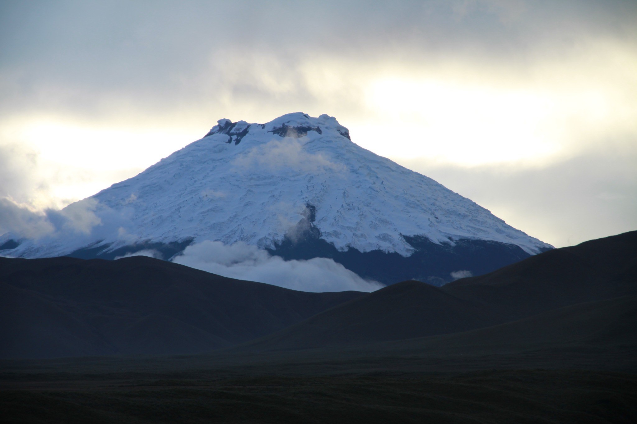 Snow-covered Cotopaxi Volcano rising above dark Andean ridges beneath a bright mountain sky