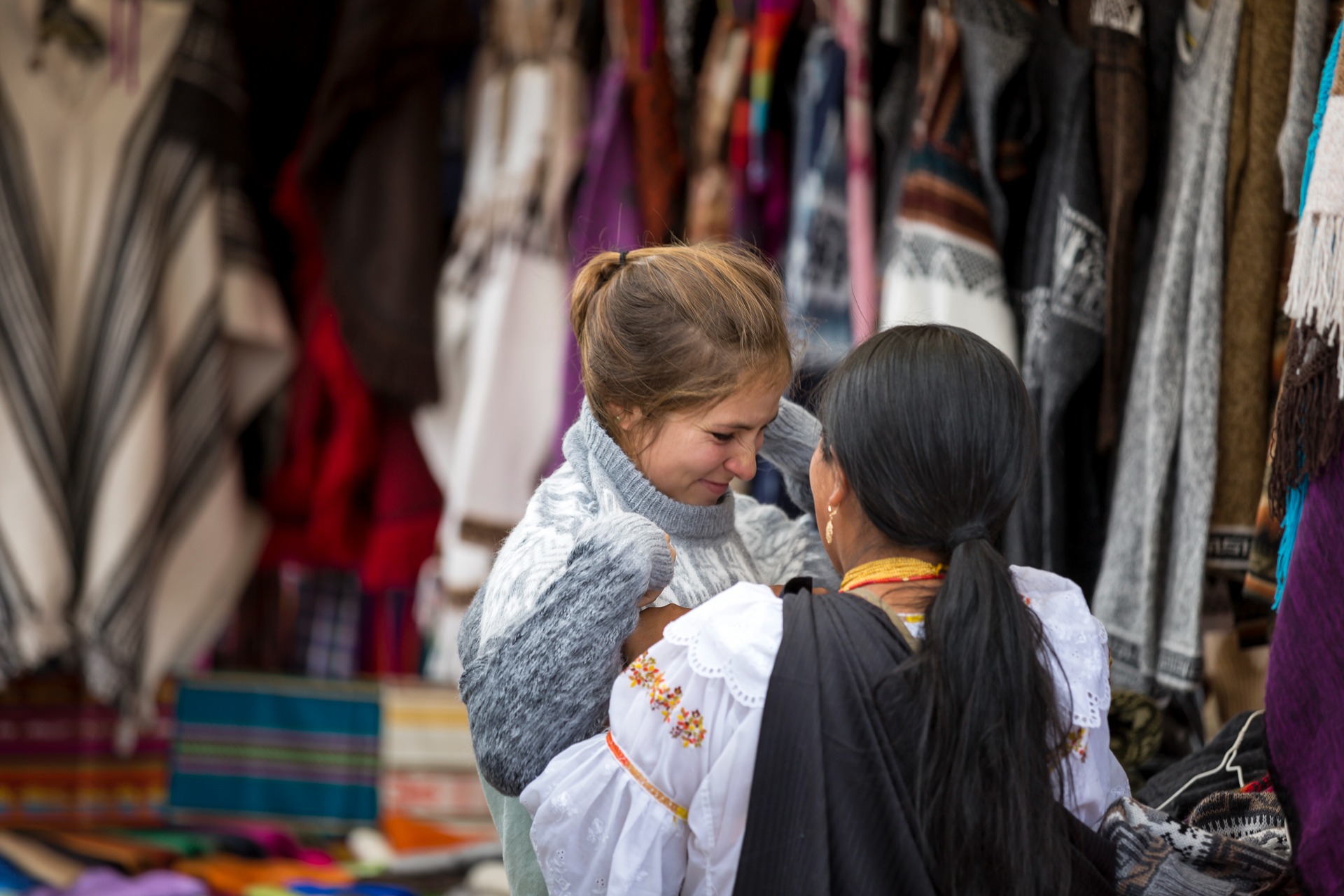 Traveler speaking with an Otavalo market vendor beside colorful woven textiles and handicrafts